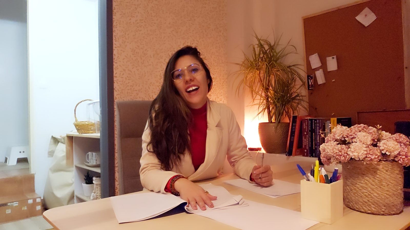 Michelle sentada à mesa de trabalho, sorrindo, com o ambiente decorado ao fundo